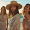 Three women in flowy floral vacation dresses standing on a sunny beach, enjoying the ocean breeze with palm trees and golden hour sunlight in the background — perfect summer outfit inspiration.