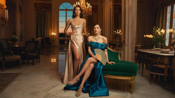 Two elegant women modeling luxurious formal and evening dresses in a grand mansion room with crystal chandeliers, marble floors, and ornate decor, showcasing satin and structured gowns in a sophisticated, high-fashion setting.