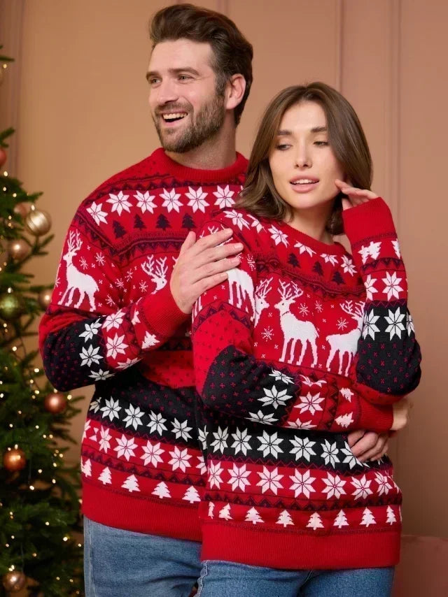 Two people wearing matching red Christmas sweaters with reindeer and snowflake patterns in front of a decorated Christmas tree.